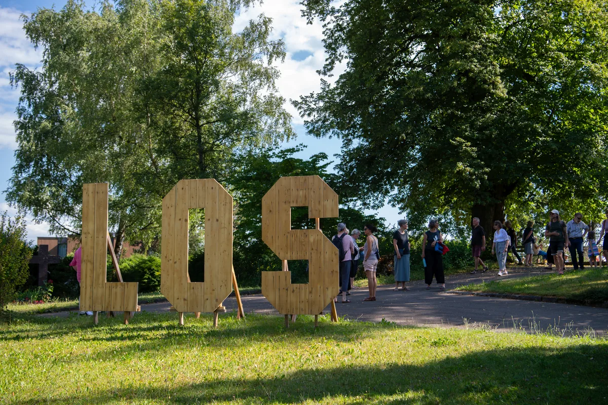 Das Wort «LOS», geschrieben mit Holzbuchstaben, daneben einige Festivalbesucher.